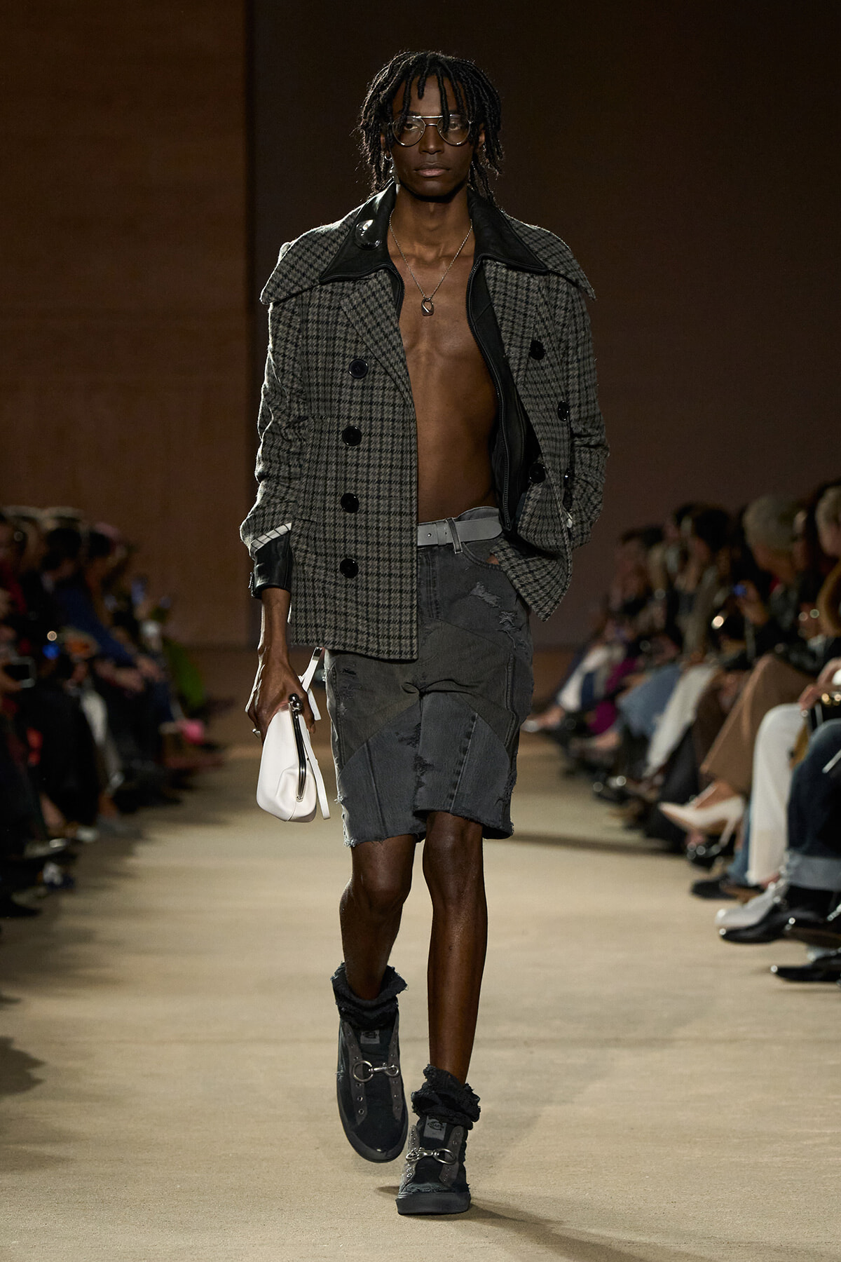 Male model walks the runway in a gray plaid oversized coat, open at the chest, paired with distressed gray shorts and dark boots with a white clutch edge accessory.