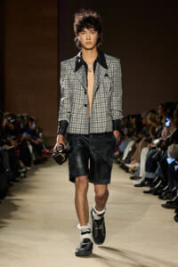 Male runway model wearing a gray plaid blazer with black lapels, black leather shorts, and fur-lined socks with sandals, carrying a small black bag.