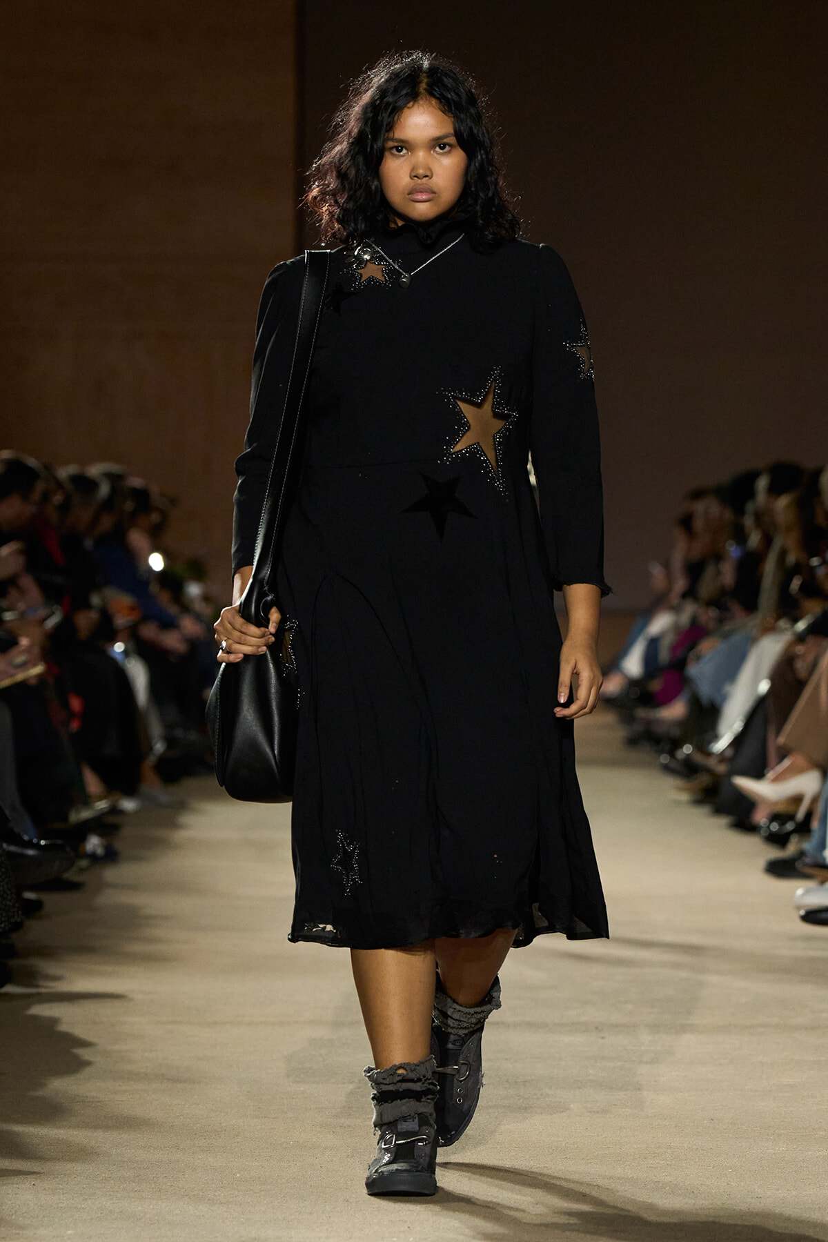 Model walks the runway in a black knee-length dress with gold and black star embellishments, carrying a black shoulder bag.