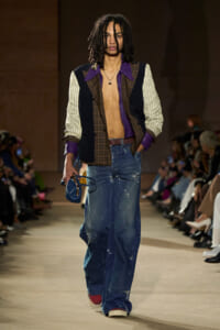 Male model walking the runway in a color-block blazer with purple lapels, open chest, and distressed wide-leg jeans, holding a blue bag.