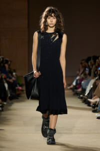 Model walking a fashion show in a sleeveless black midi dress with subtle sparkle, carrying a large black shoulder bag.