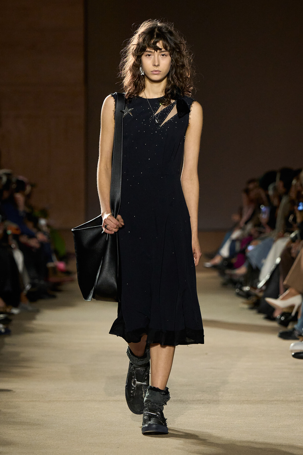Model walking a fashion show in a sleeveless black midi dress with subtle sparkle, carrying a large black shoulder bag.