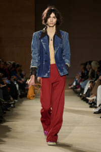 Model walking the runway in a distressed blue denim blazer, red wide-leg pants, purple sneakers, and a small tan handbag.