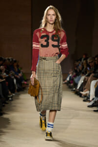 Model walking a runway in a red-pink varsity-style top with the number 39, a beige-and-black plaid midi skirt, white socks, yellow sneakers, and a tan fringe handbag.