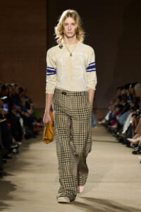 Female model walking a fashion runway in a cream sweater with blue striped sleeves and a prominent '19' on the front, paired with plaid wide-leg pants, carrying a yellow clutch.
