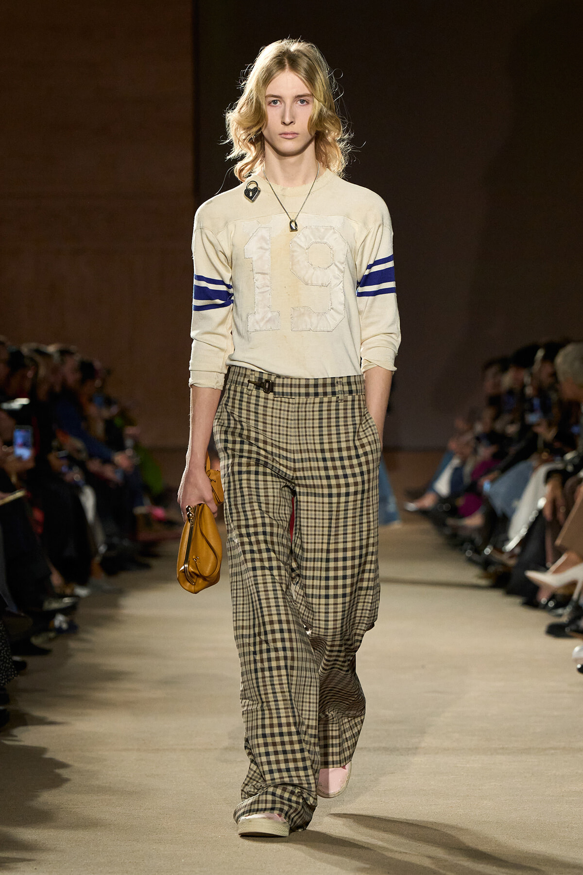 Female model walking a fashion runway in a cream sweater with blue striped sleeves and a prominent '19' on the front, paired with plaid wide-leg pants, carrying a yellow clutch.