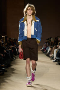Male runway model wearing a blue and white color-blocked varsity jacket, brown shorts, and bright purple high-top sneakers; he holds a red clutch and walks the catwalk.