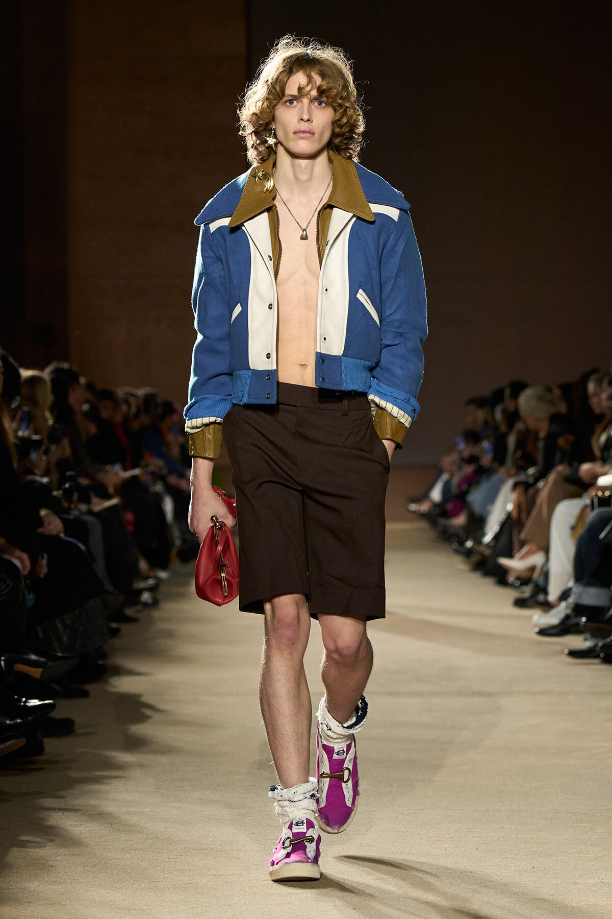 Male runway model wearing a blue and white color-blocked varsity jacket, brown shorts, and bright purple high-top sneakers; he holds a red clutch and walks the catwalk.