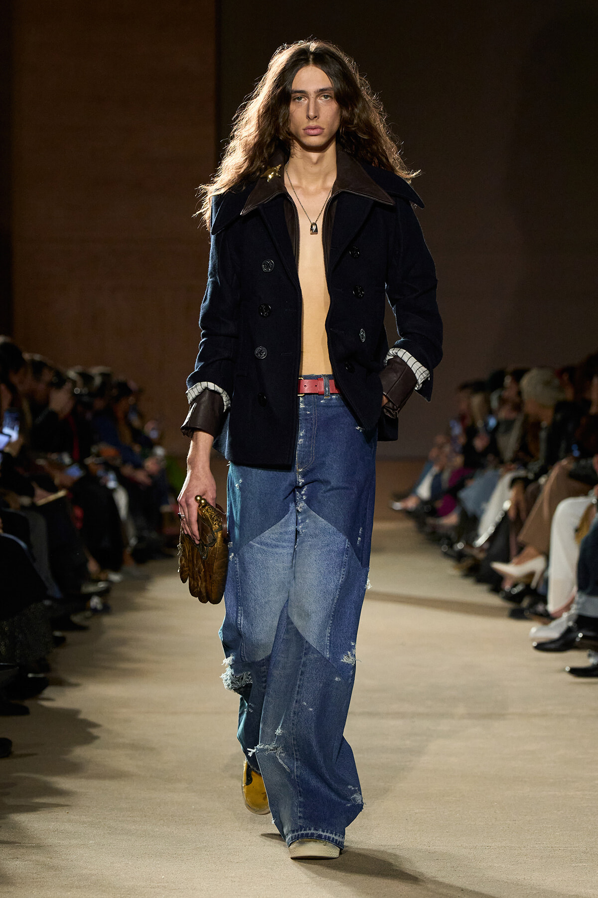 Male model walking a runway in a dark navy double-breasted coat, open to reveal a bare chest, paired with patchwork denim jeans and a red belt, holding a brown clutch.