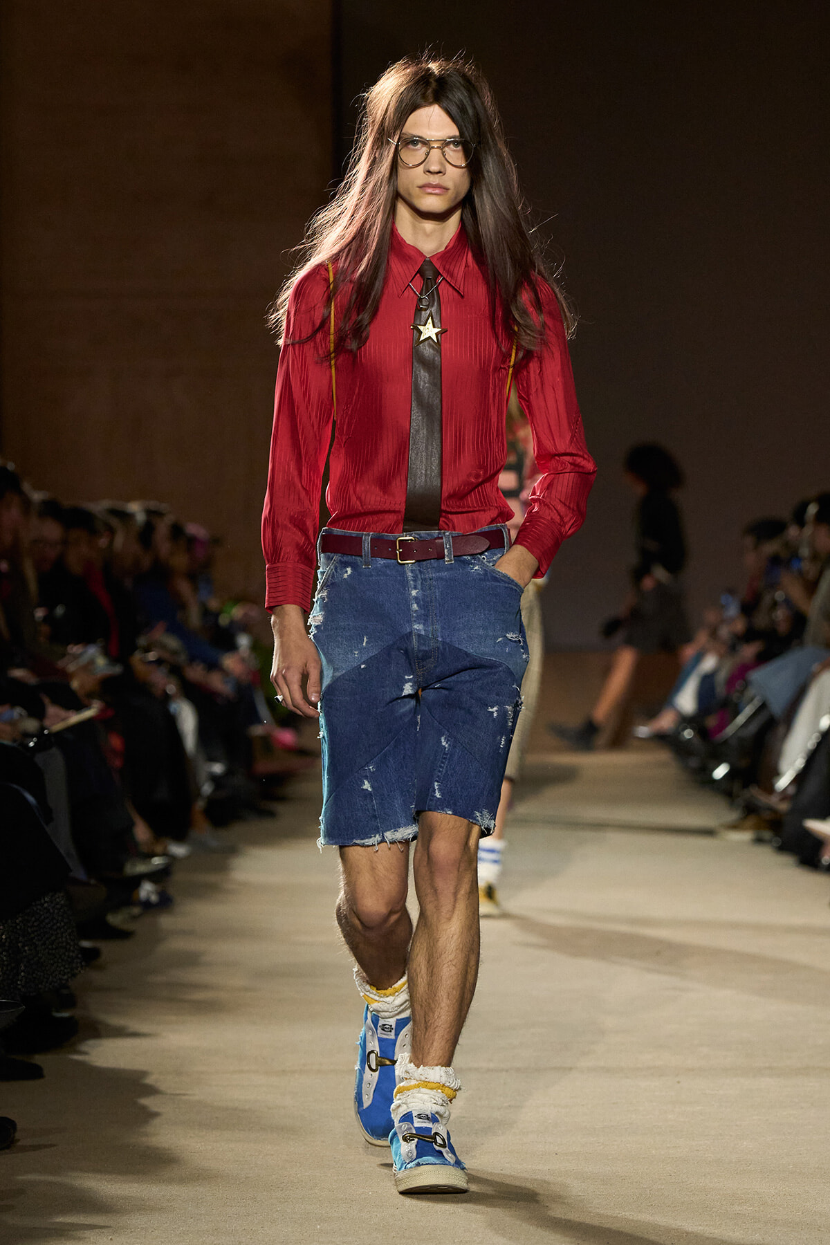 Model walking a fashion show runway in a red shirt with a black tie, suspenders, and distressed denim shorts, paired with blue sneakers.