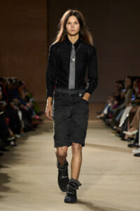 Runway model in an all-black outfit: black shirt, gray tie with a star pendant, distressed knee-length denim skirt, and fur-trimmed boots.