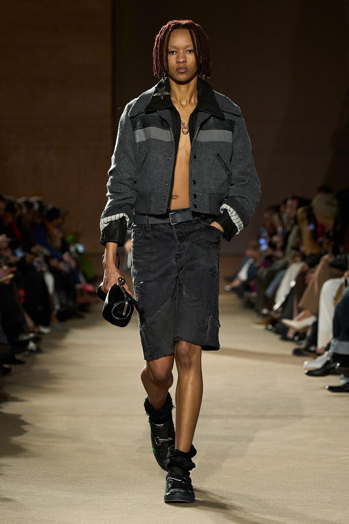 Model on a runway wearing a gray cropped jacket, a distressed dark denim skirt with a belt, and green-trim boots, carrying a small black purse.