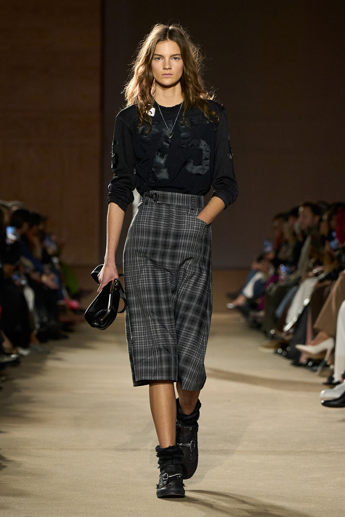 Model on a runway wearing a black patched top, gray plaid wide-leg culottes, and black fur-trim boots, carrying a small black bag.