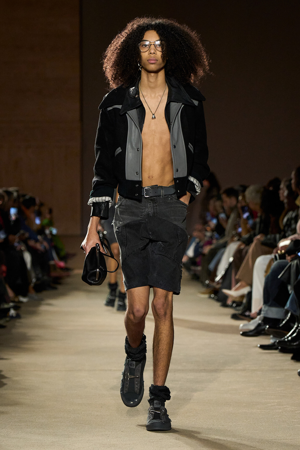 Male model walks the runway in a two-tone black/gray varsity-style jacket, open to reveal bare chest, paired with a black denim skirt and boots, accessories include glasses and a pendant necklace.