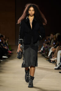 Female model walks a runway in a black blazer, dark midi skirt, layered necklaces, and chunky sneakers with scrunched socks, audience in the background.