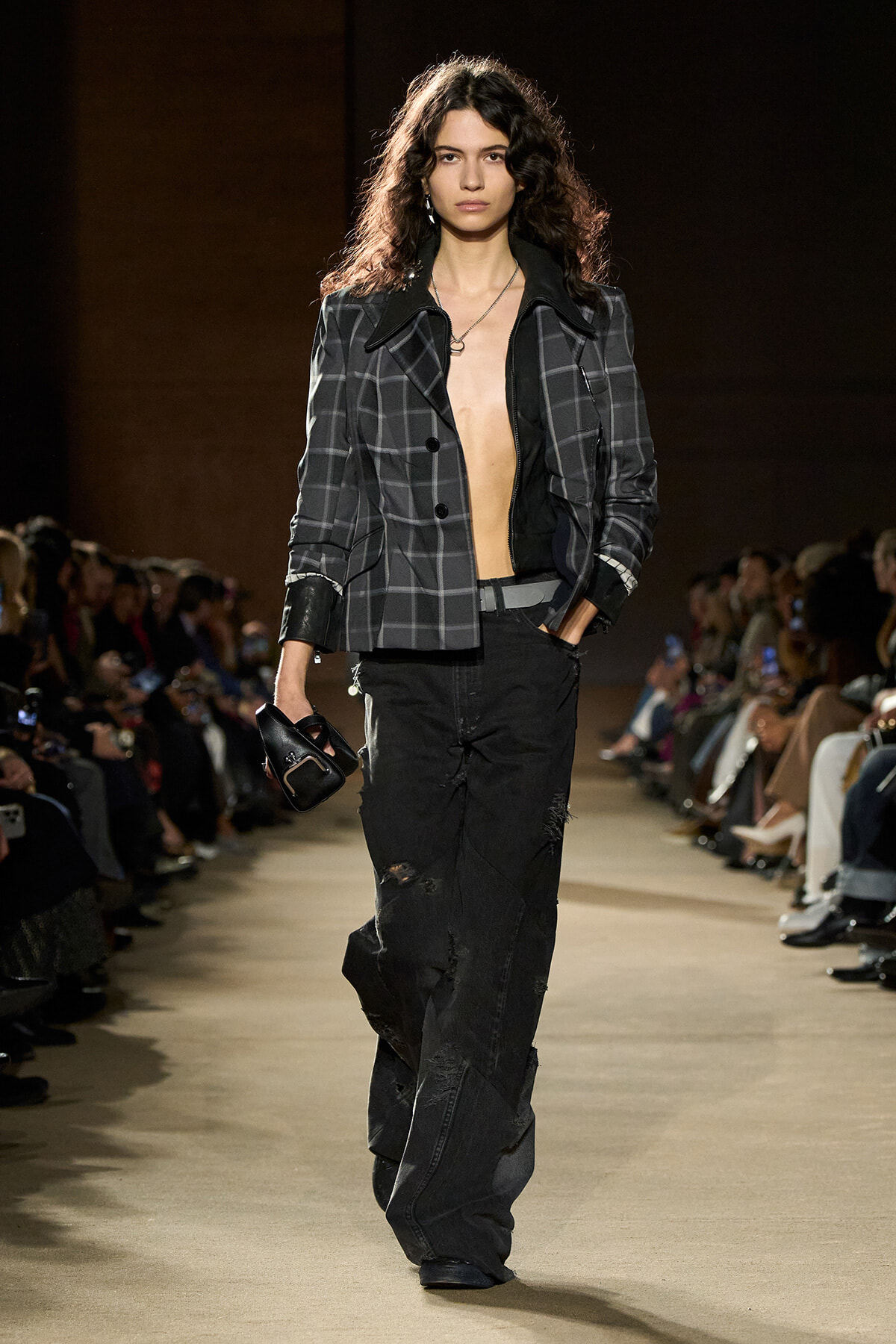 Model walking the runway in a gray plaid blazer over a bare chest, paired with distressed black jeans and a small black handbag