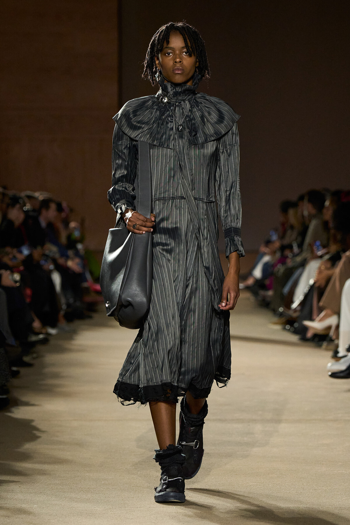Model walking runway in a long gray pinstripe dress with a large ruffled collar, carrying a gray shoulder bag and wearing chunky boots.