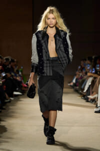 Model walking the runway in a patchwork blazer, distressed black skirt, combat boots, and a small black clutch. Audience visible in the background.