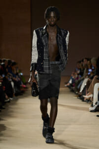Male model on a runway wearing a celestial-print blazer over a plaid jacket, open chest, black distressed shorts, chunky boots, and a small black clutch.