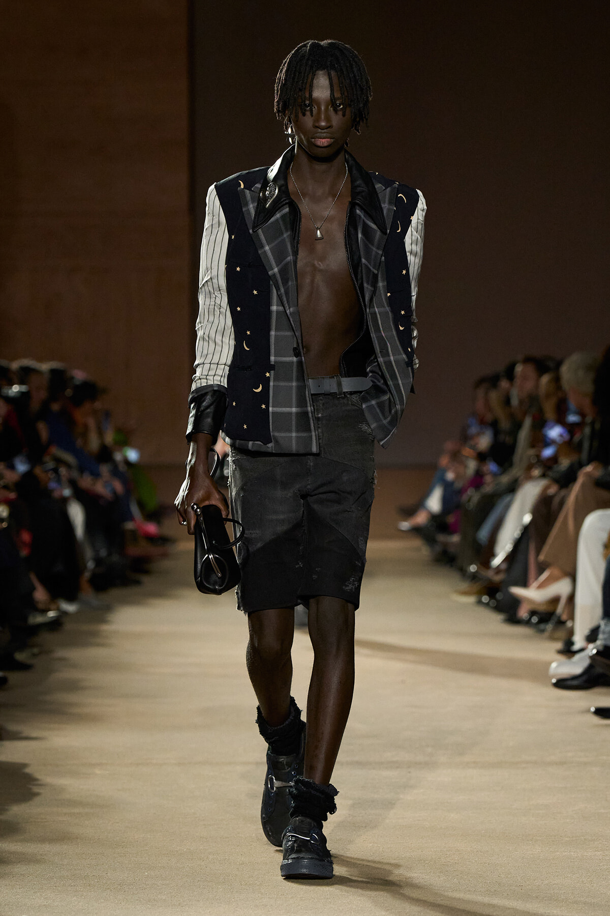 Male model on a runway wearing a celestial-print blazer over a plaid jacket, open chest, black distressed shorts, chunky boots, and a small black clutch.