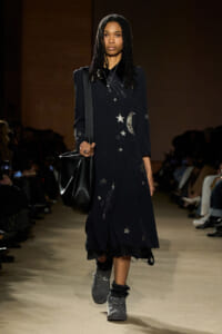 Model walks a runway in a navy dress with silver stars and a crescent moon embroidery, carrying a black shoulder bag and wearing gray boots.
