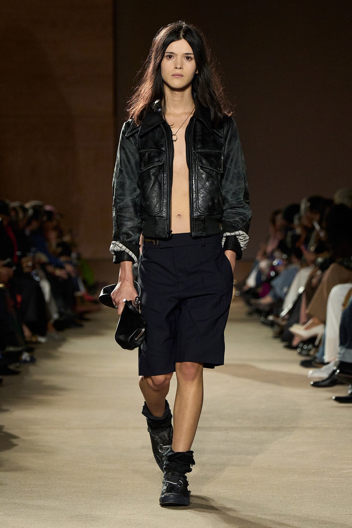 Model walks a runway in a black leather bomber jacket, navy knee-length shorts, and rugged boots, carrying a small black bag.
