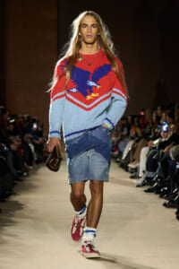 Male model on a runway wearing a red‑and‑blue knit sweater with a bird motif, paired with patchwork denim shorts.