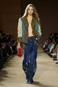 Model walking runway in patchwork jacket, distressed jeans, and a red clutch on a beige catwalk.