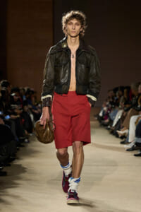 Male model walks a runway wearing a black leather jacket, red knee-length shorts, and bold sneakers with socks; holding a brown bag.