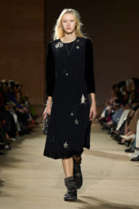 Model walking a fashion runway in a black midi dress with velvet sleeves and star embroidery, holding a small bag.