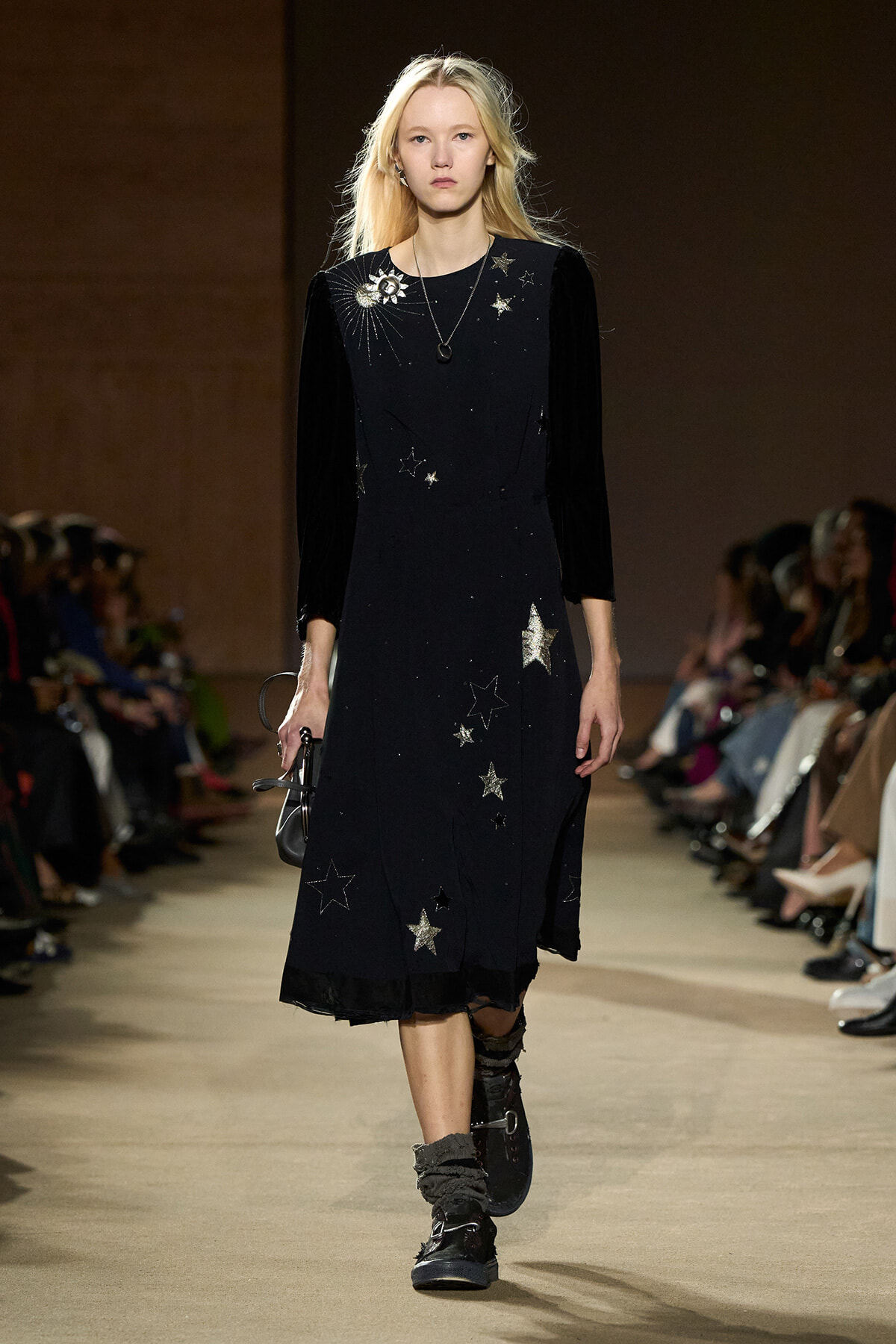 Model walking a fashion runway in a black midi dress with velvet sleeves and star embroidery, holding a small bag.