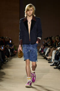 Male model walks the runway in a navy blazer with burgundy lapels, no shirt, distressed denim shorts, pink sneakers, and a brown clutch.