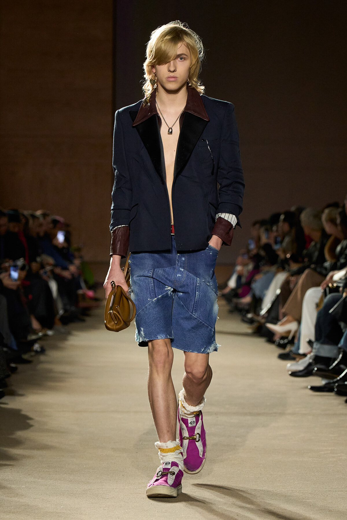 Male model walks the runway in a navy blazer with burgundy lapels, no shirt, distressed denim shorts, pink sneakers, and a brown clutch.