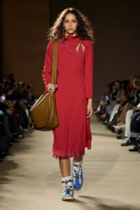 Model walking down a fashion show runway in a red, long-sleeve dress with decorative cutouts, carrying a large brown shoulder bag and wearing blue sneakers with white fur socks.