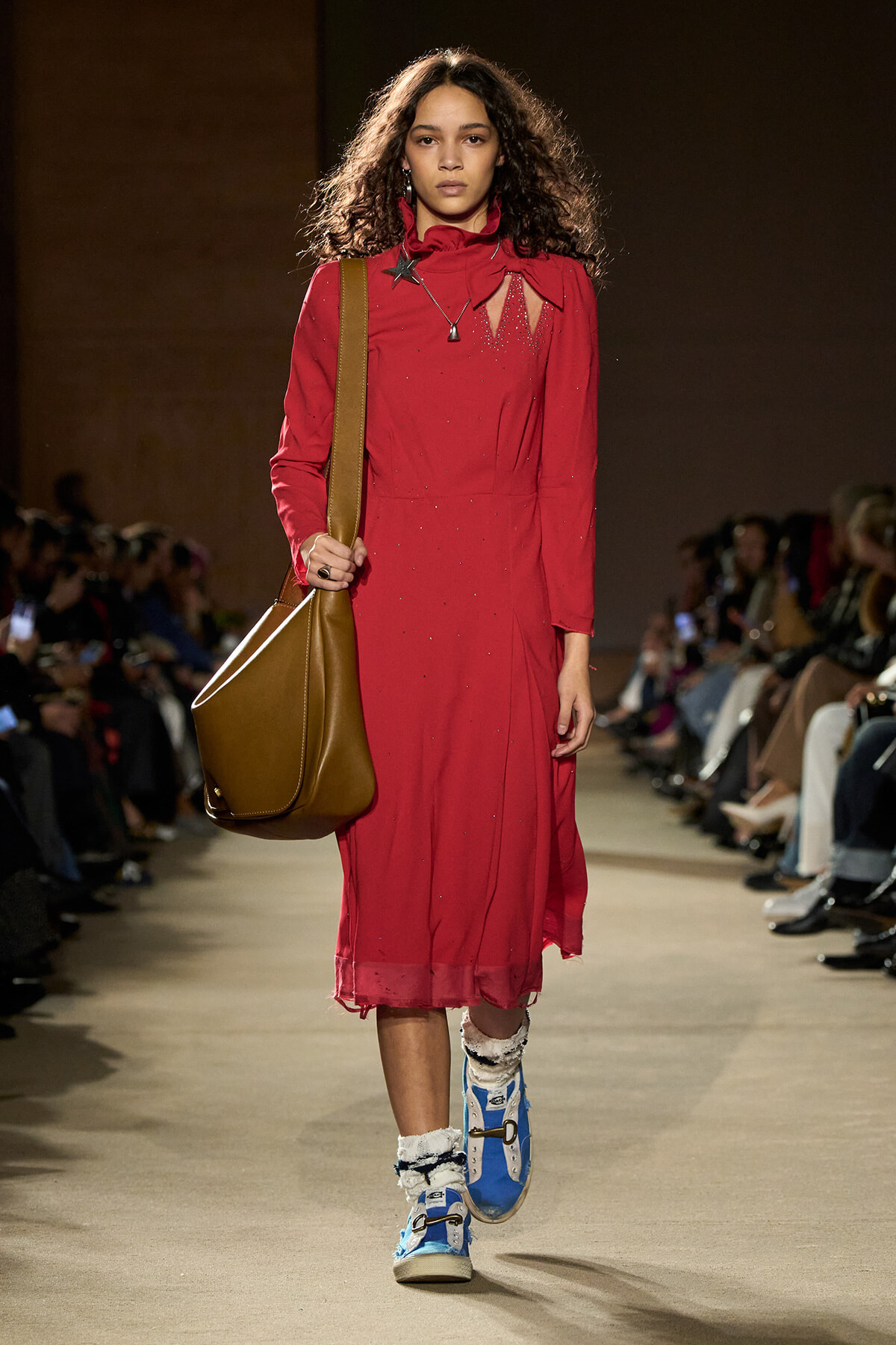 Model walking down a fashion show runway in a red, long-sleeve dress with decorative cutouts, carrying a large brown shoulder bag and wearing blue sneakers with white fur socks.