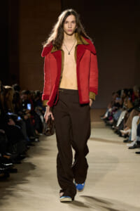 Male model walking runway in a red shearling jacket with brown cuffs, no shirt, brown wide-leg trousers, blue sneakers, and a small brown bag.