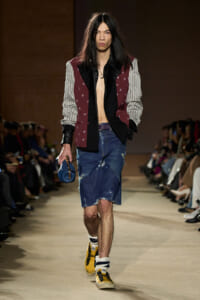 Runway model with long dark hair wearing a burgundy star-pattern blazer, striped sleeves, no shirt, distressed denim shorts, yellow sneakers, and a blue clutch.