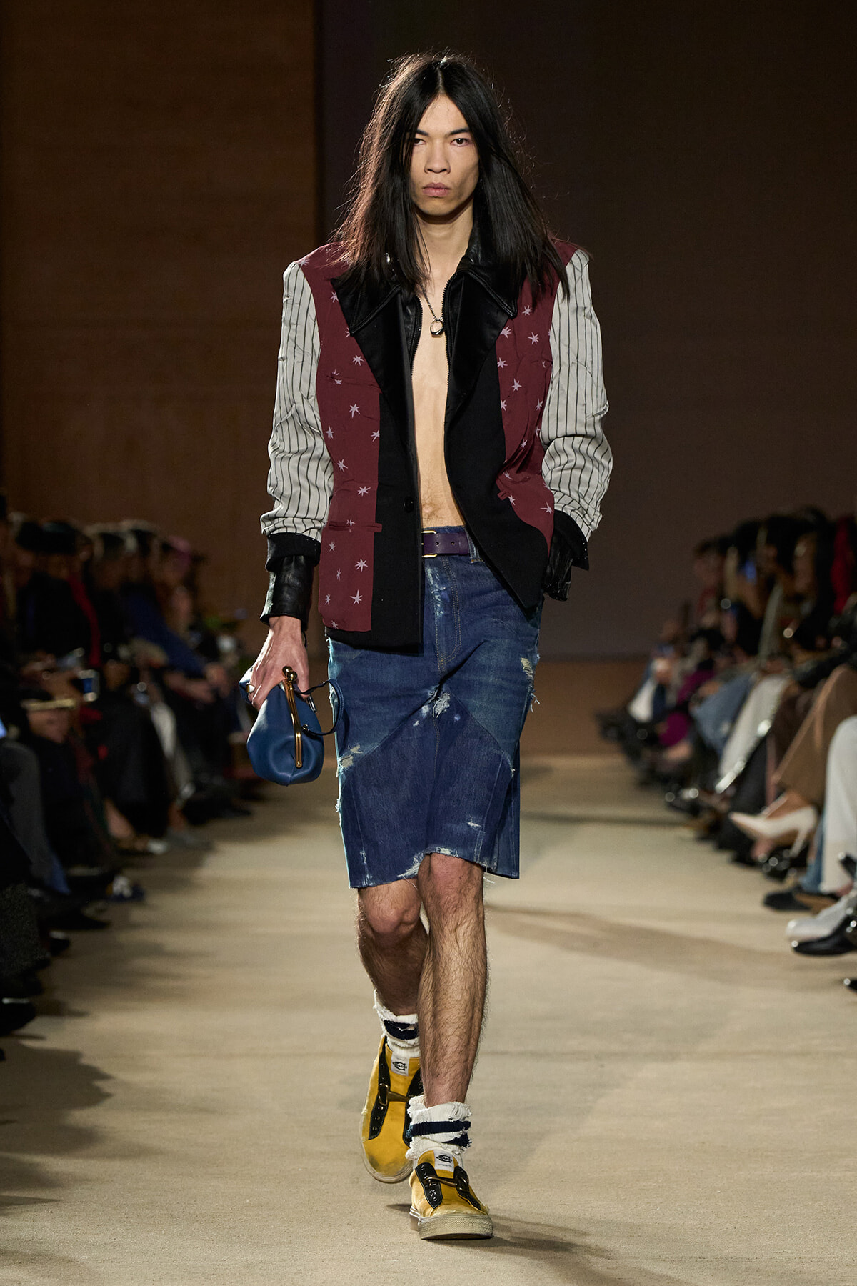 Runway model with long dark hair wearing a burgundy star-pattern blazer, striped sleeves, no shirt, distressed denim shorts, yellow sneakers, and a blue clutch.