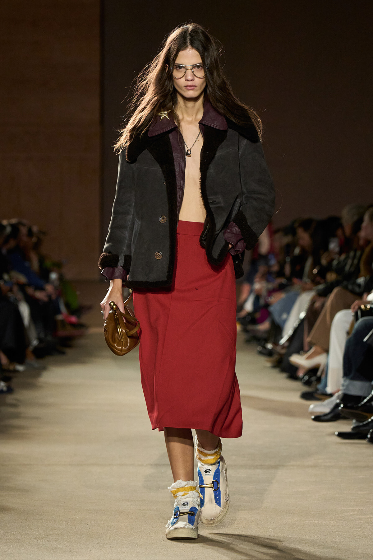 Model walking a runway in a dark oversized jacket, red midi skirt, and blue‑yellow sneakers, holding a brown handbag.