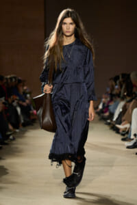 Female model walking a fashion runway in a navy pinstripe midi dress with a frayed hem, carrying a large brown leather shoulder bag and wearing black boots.