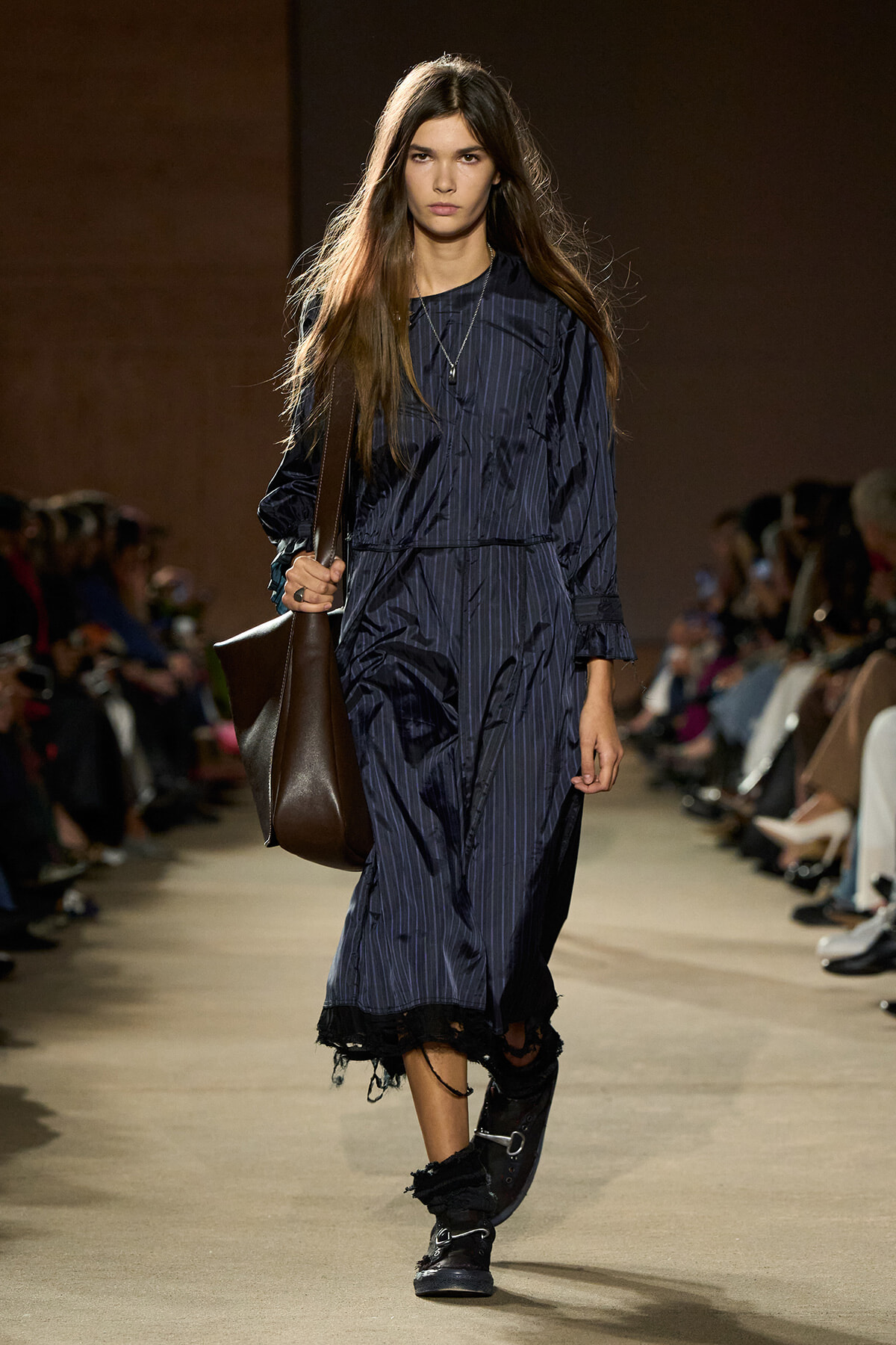 Female model walking a fashion runway in a navy pinstripe midi dress with a frayed hem, carrying a large brown leather shoulder bag and wearing black boots.