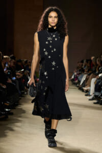 Model walks the runway in a black sleeveless midi dress adorned with silver stars, holding a plaid clutch.