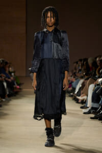 Model on a runway wearing a dark navy midi dress with layered panels and puff sleeves, black boots.