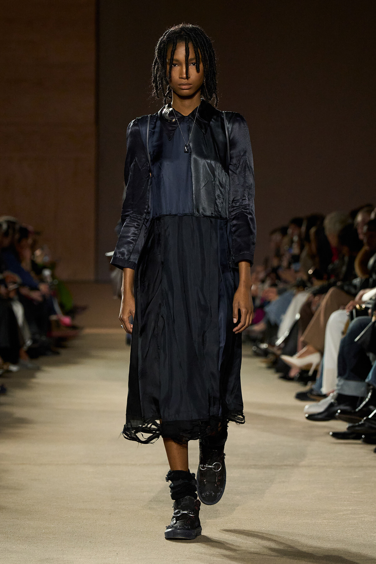 Model on a runway wearing a dark navy midi dress with layered panels and puff sleeves, black boots.