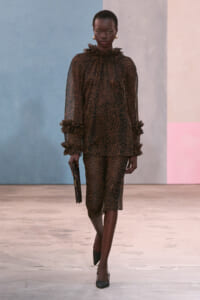 Model walking a runway in a brown leopard-print sheer blouse with ruffled collar and cuffs, matching midi skirt, black clutch, and dark heels.