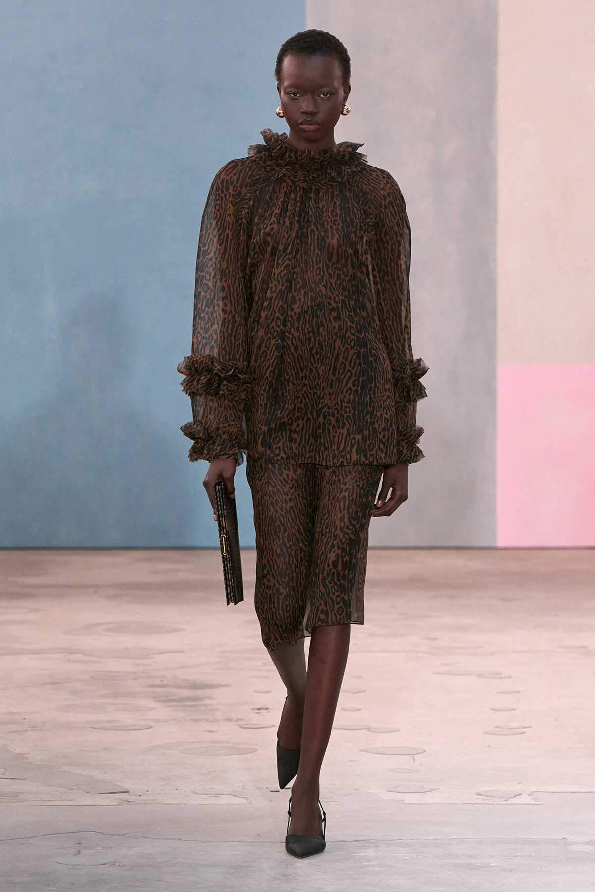 Model walking a runway in a brown leopard-print sheer blouse with ruffled collar and cuffs, matching midi skirt, black clutch, and dark heels.