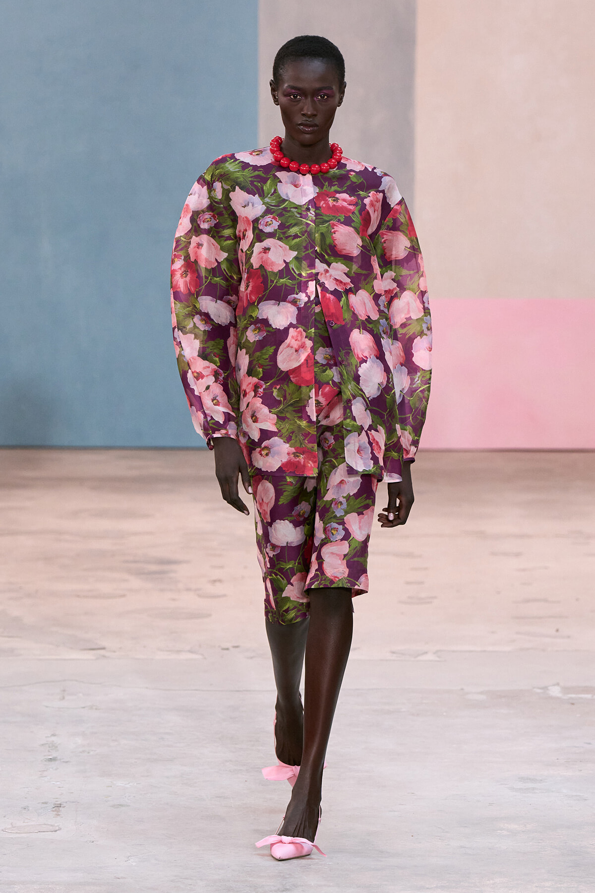 Female model walking a runway in a purple floral suit with pink flowers, a bold red beaded necklace, and pink bow flats.