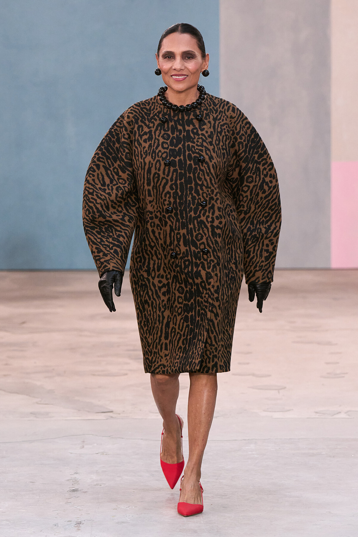 Female model wearing a brown and black leopard-print knee-length coat with oversized sleeves, black gloves, and bold black bead jewelry walking on a runway; bright red pointed heels add a pop of color.