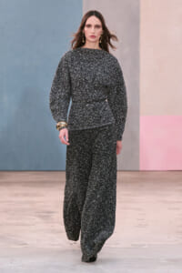 Model walks the runway in a textured gray-black tweed matching set with a belted waist and wide-leg pants, accessorized with gold earrings and bracelets.