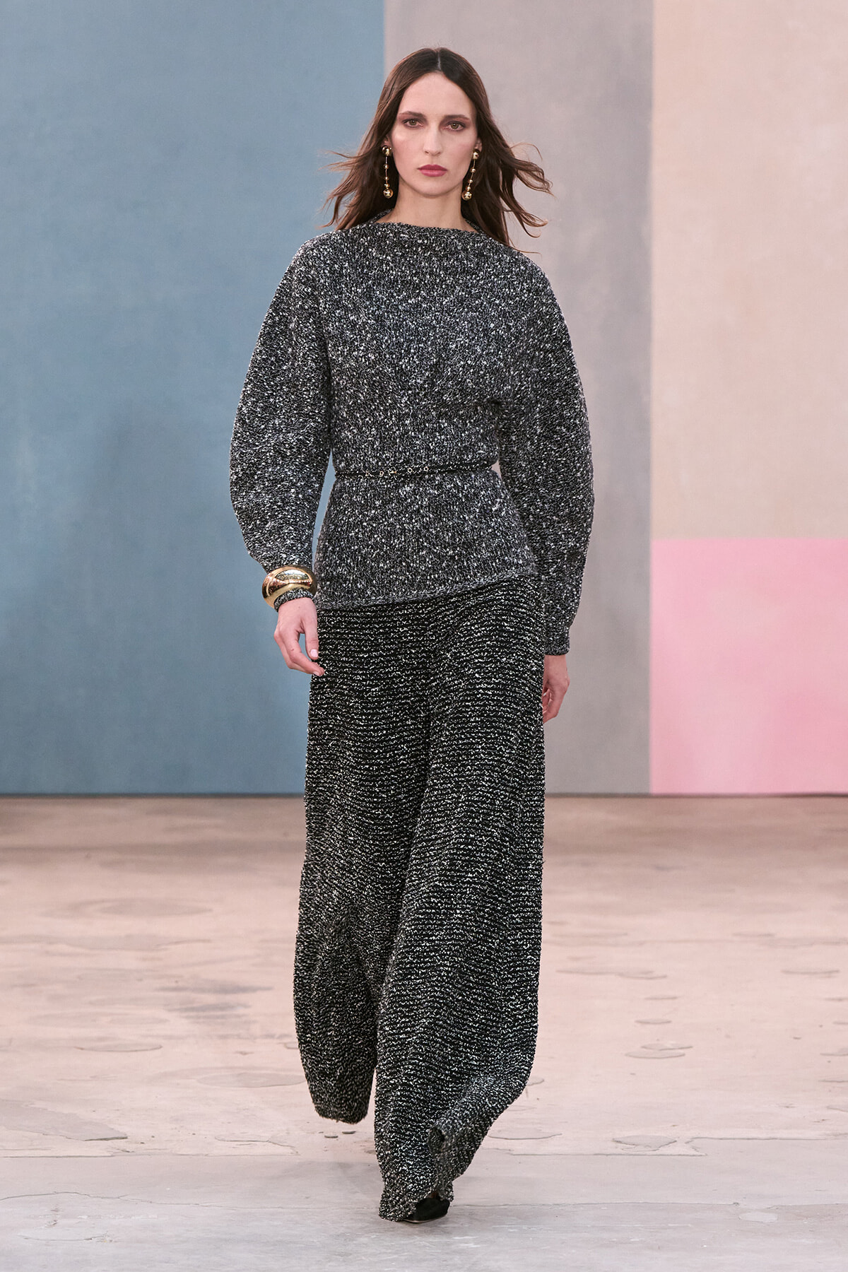 Model walks the runway in a textured gray-black tweed matching set with a belted waist and wide-leg pants, accessorized with gold earrings and bracelets.